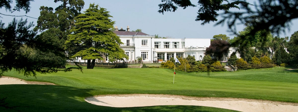 A view of Nutley House which is the name of the clubhouse in the far distance the house is two storeys high white in colour and neo Georgian in style and there is a bunker with a pinhole in the forefront of the picture
