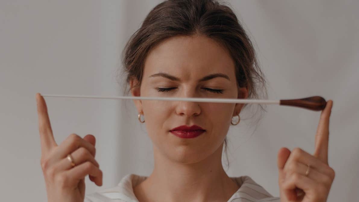 A woman with closed eyes is holding up a baton in front of her face between two index fingers