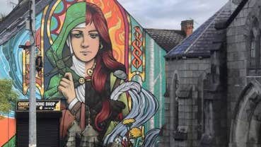 Cruinniú na nÓg 2026 Walking With Legends Live, a group of people on a pavement looking up at the end of building covered in a mural of a woman.