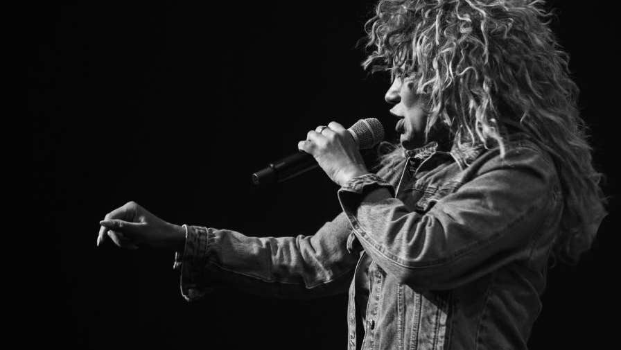 Side view of woman with big hair singing into a mic, against dark, plain background.