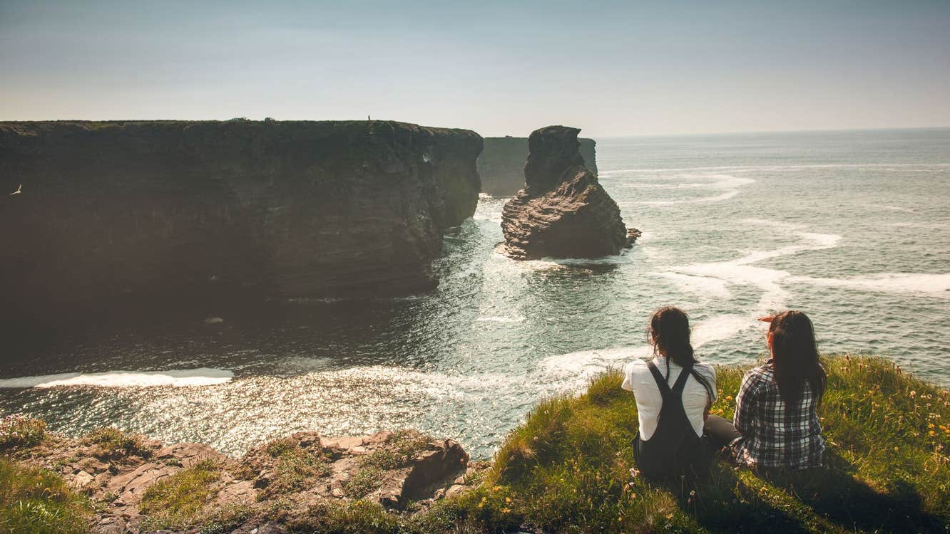 Two ladies sit on a grass verge on the coast looking across at cliffs in the distance