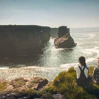 Two ladies sit on a grass verge on the coast looking across at cliffs in the distance