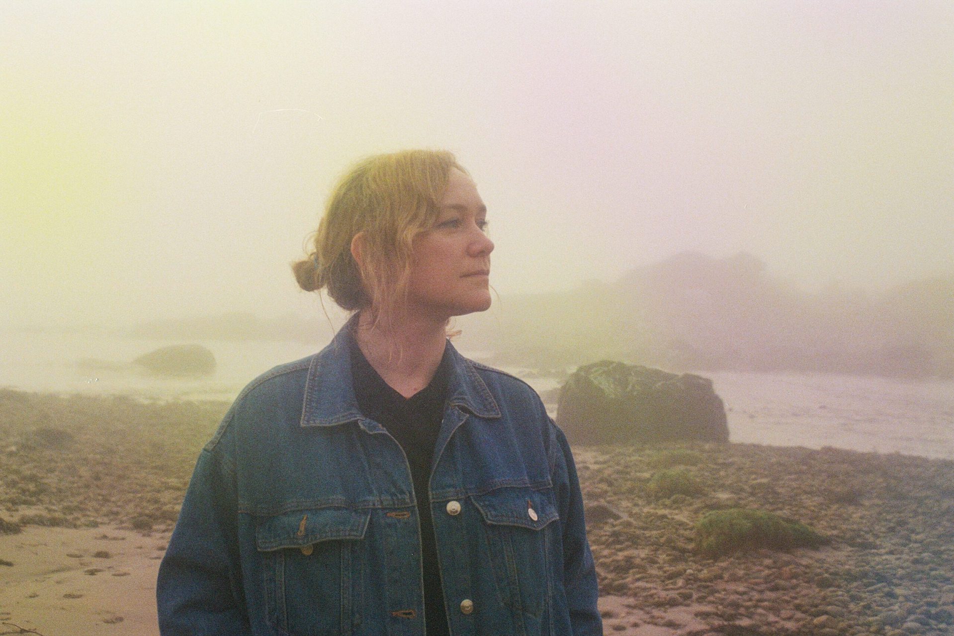 A woman in denim jacket is outdoors, looking away to her left looking reflective, against rocky shoreline blurred by mist.