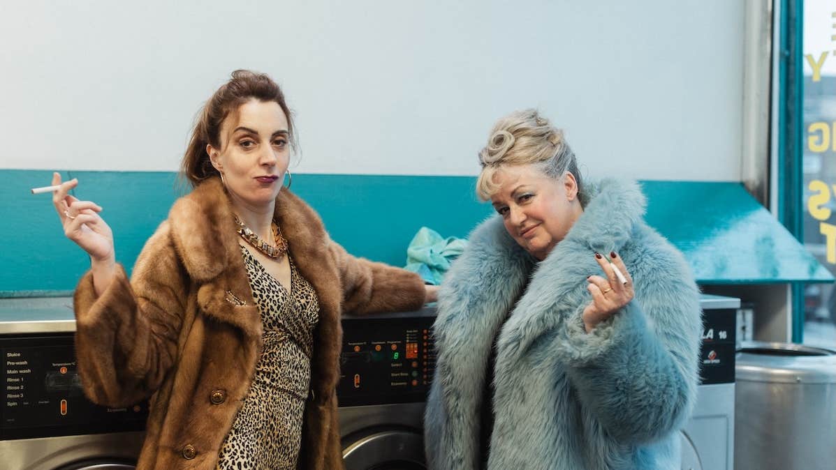 2 women in fur coats are holding cigarettes, leaning against washing machines in a launderette.