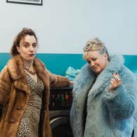 2 women in fur coats are holding cigarettes, leaning against washing machines in a launderette.