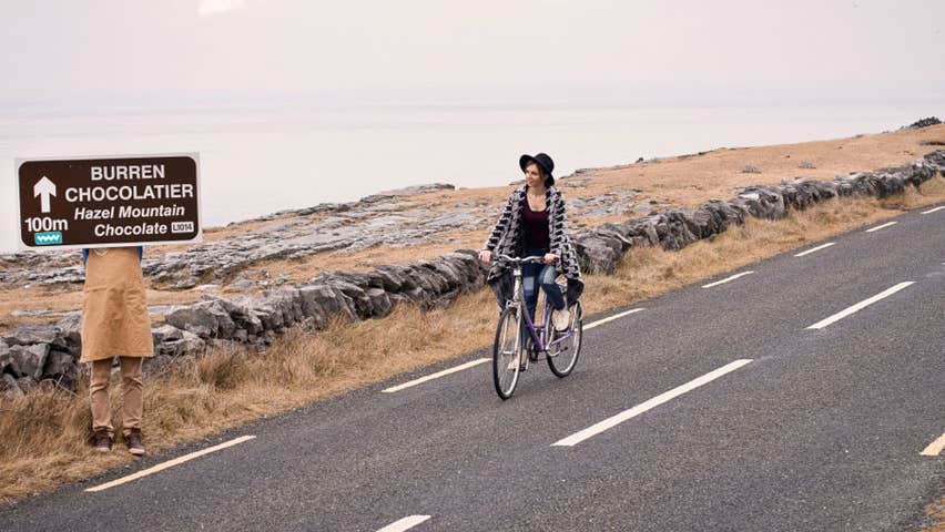 A woman cycling her bicycle along a coastal road while passing by a Burren Chocolatier road sign