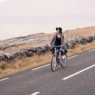 A woman cycling her bicycle along a coastal road while passing by a Burren Chocolatier road sign