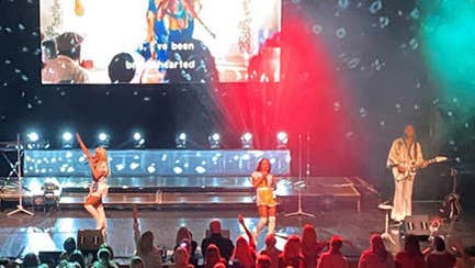 View of a stage looking over the audience with 2 women singing and 2 men playing instruments with large screen at the back of the stage.