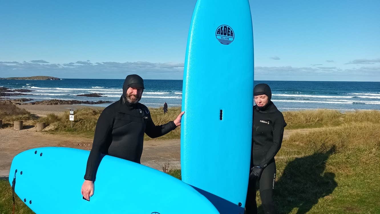 Two people standing on grass in wetsuits holding blue surf boards by a beach