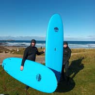Two people standing on grass in wetsuits holding blue surf boards by a beach