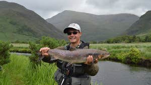 Man holding a fish, standing on the riverbank with mountains in the background