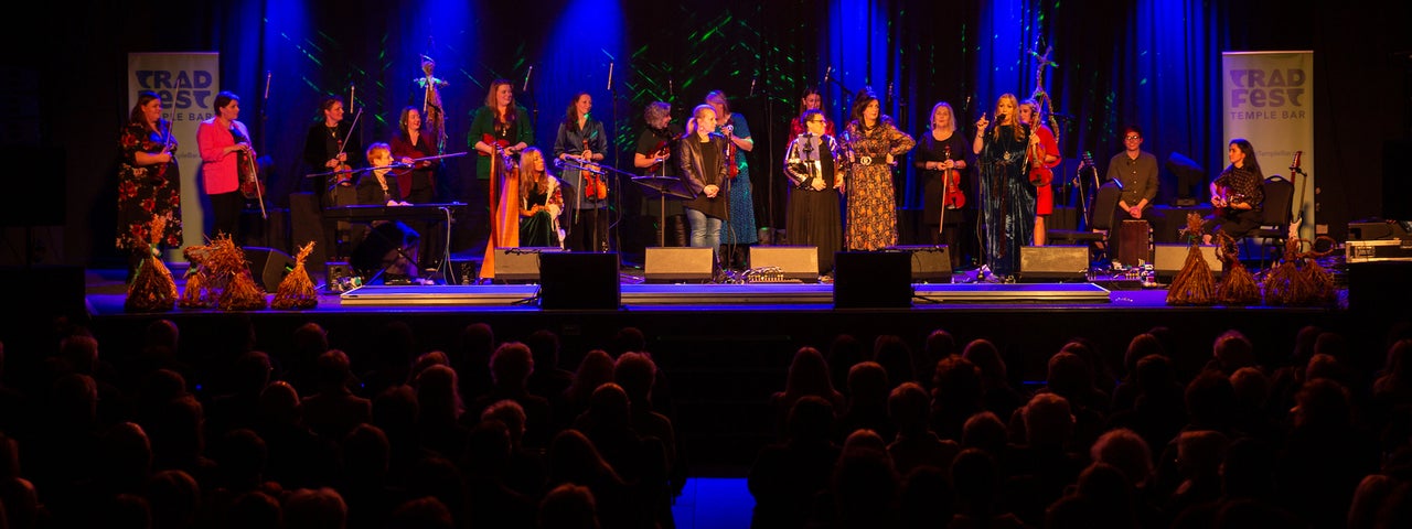 A stage seen from a distance with large group of women in 2 rows, singing and playing musical  instruments.