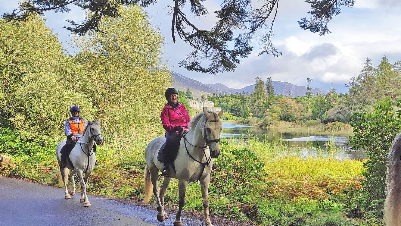 Connemara Equestrian Escapes view of two people on horseback with a lake and trees and mountains behind