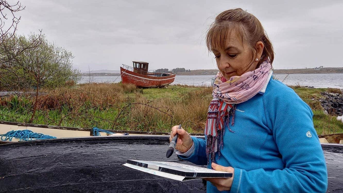 An artist with a pad and paintbrush in hand with an old boat in the background