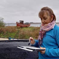 An artist with a pad and paintbrush in hand with an old boat in the background