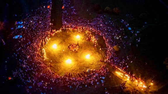 Viewed from above at night, a large crowd gathered around a large circle containing 4 fires.