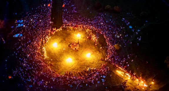 Viewed from above at night, a large crowd gathered around a large circle containing 4 fires.
