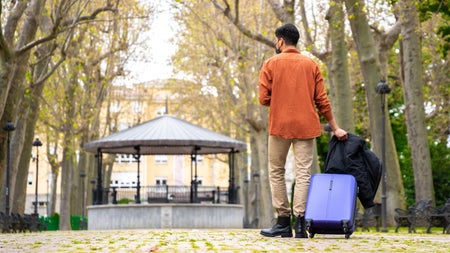 A man standing with a suitcase and jacket in a city park