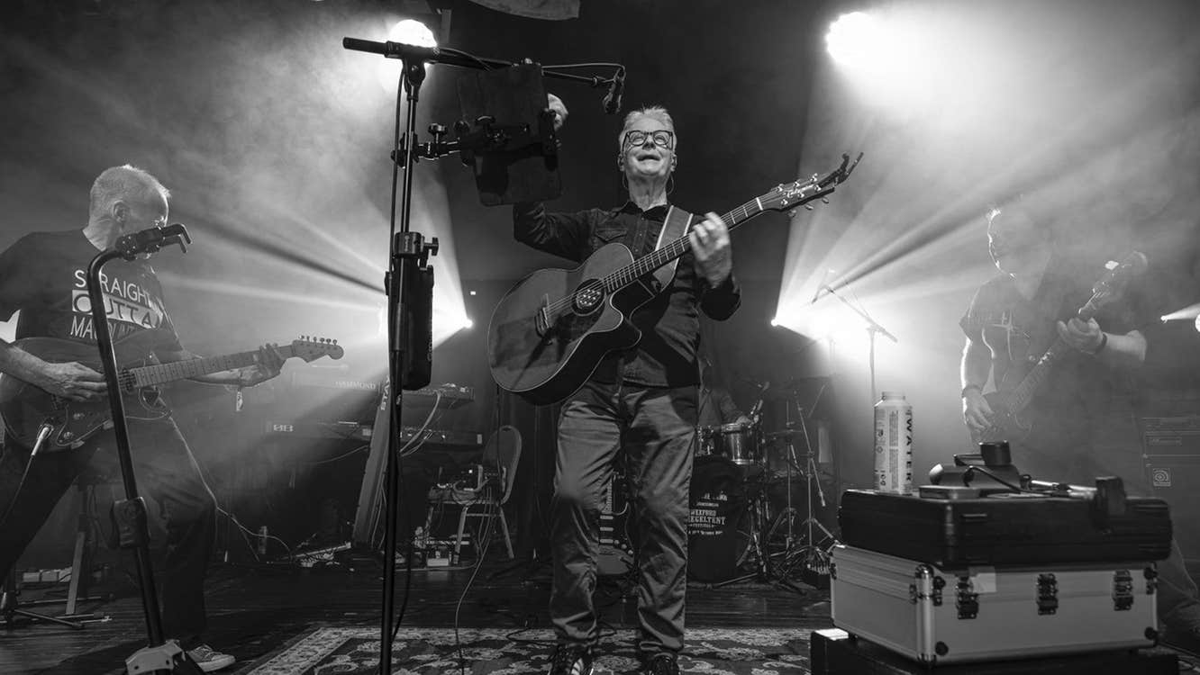 Black and white photo of man with guitar on a stage with 2 bright lights shining outwards either side of him.