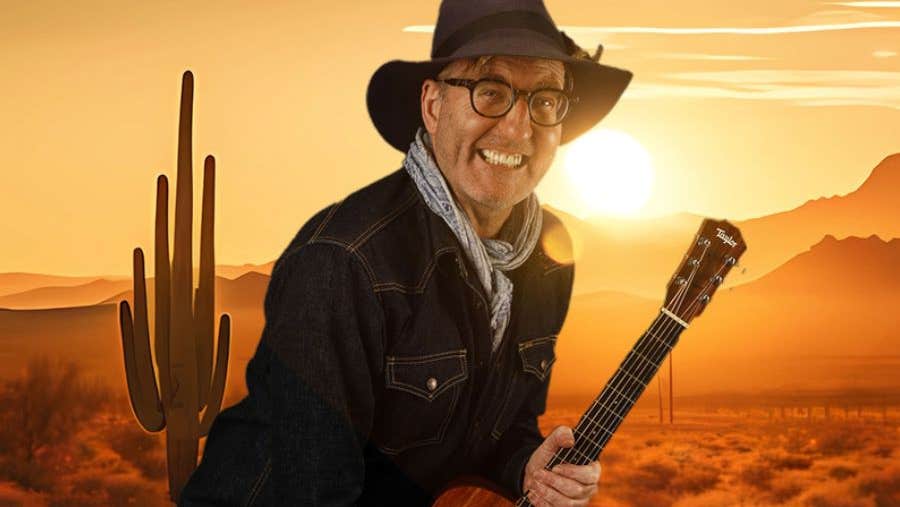 A grinning man in dark hat and jacket is clutching a guitar against backdrop of a desert with yellow and orange setting sun.