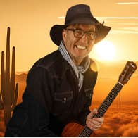 A grinning man in dark hat and jacket is clutching a guitar against backdrop of a desert with yellow and orange setting sun.