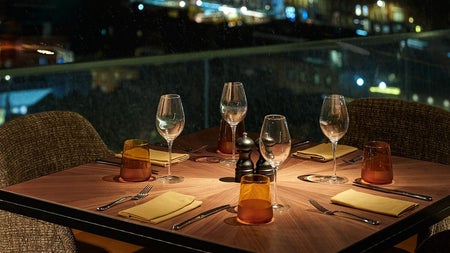 A rooftop dining table set with wine glasses and cutlery overlooking the city skyline at night