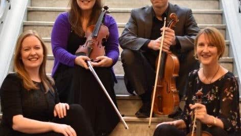 4 smiling people are seated on stone stairs each holding string instruments