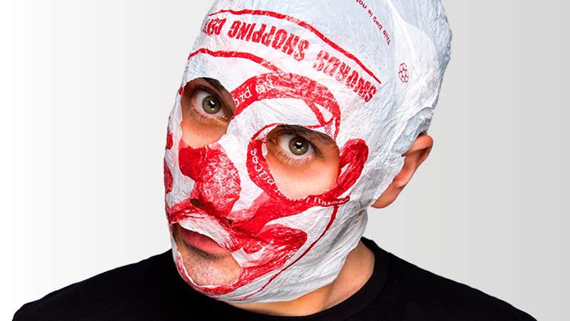 The face of a man is covered in a mask made from a white and red carrier bag, with eye and mouth holes cut away.
