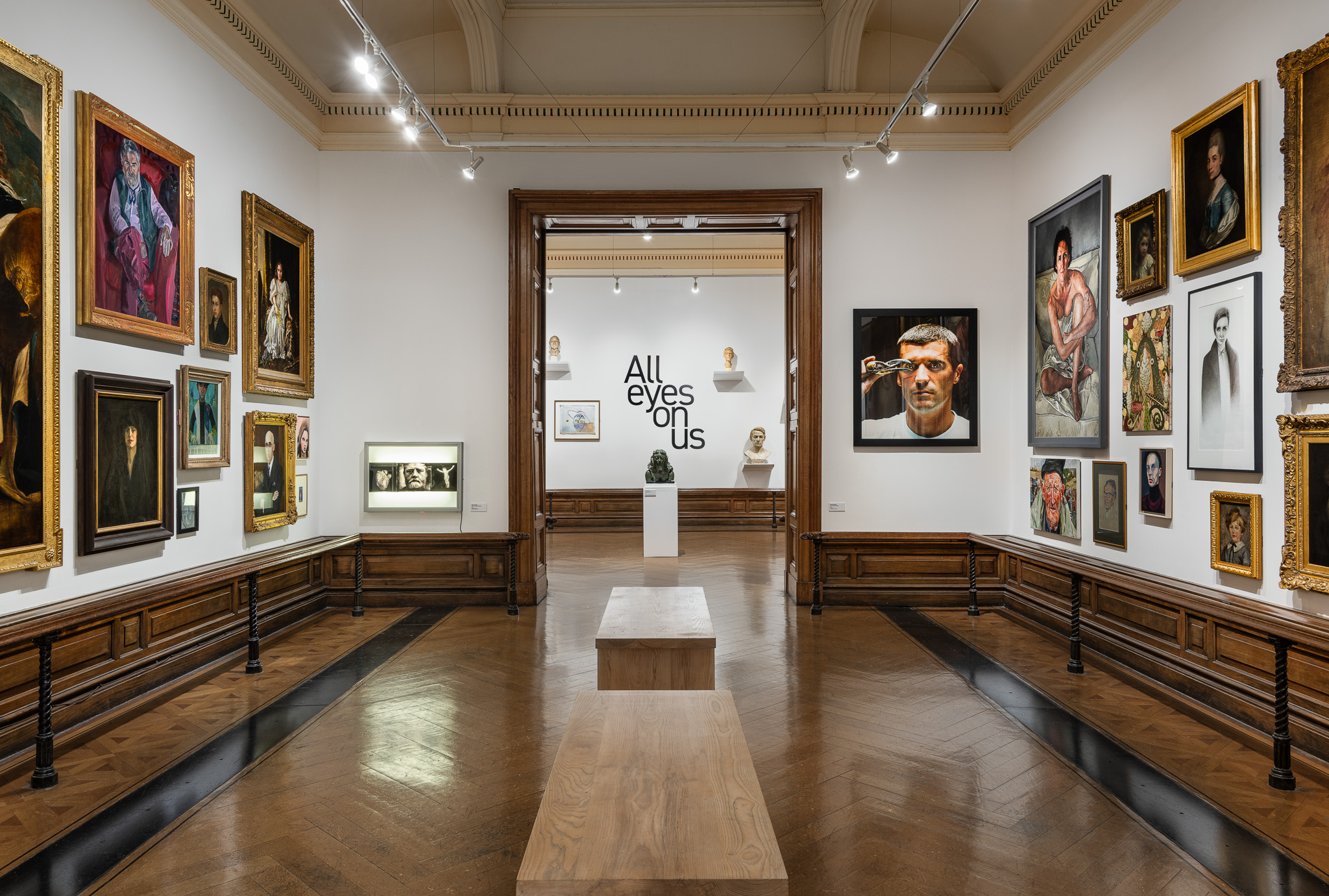 View of an art gallery with dark wooden floors, pale walls and various sizes of art hanging on the walls with view through doorway into another exhibition room.