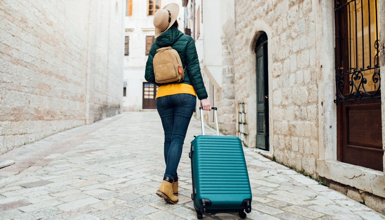 A girl looking up wearing a hat pulling her suitcase through a city street