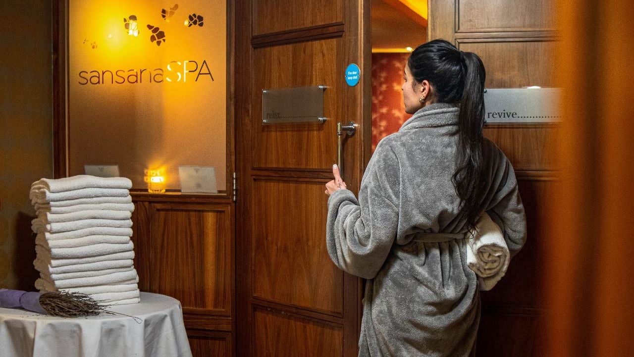 Person in a grey dressing gown holding a towel entering a room with large wooden doors