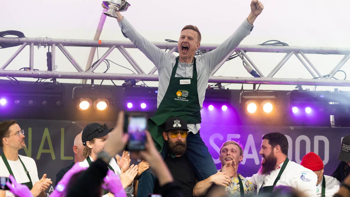 A man in black apron is celebrating with arms raised up, on the shoulders of other people with temporary event stage behind.
