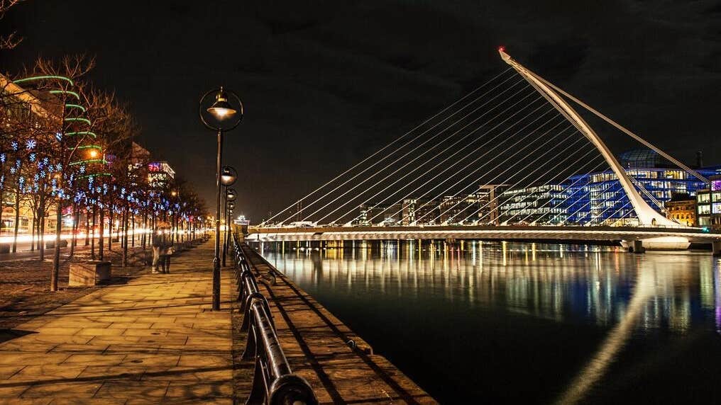 A view down alongside a river bank in the city at night with bridge lit up and buildings in the distance.