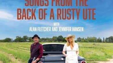 A smiling man and woman leaning against the boot of a big old car on the edge of a field.