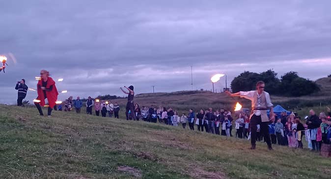 A large group of people in the distance on a hill at dusk, some holding lit flames.