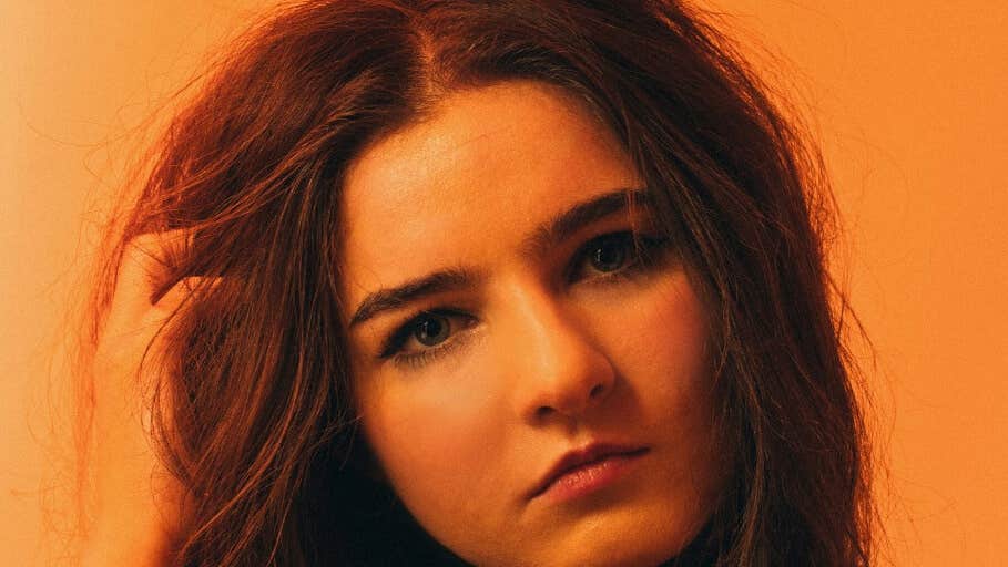 Brigit x District Muireann Bradley - headshot of Solomon woman with hand in her hair on on side, against orange background.