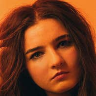 Brigit x District Muireann Bradley - headshot of Solomon woman with hand in her hair on on side, against orange background.