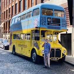 Parked blue and yellow bus with a man drinking from a takeaway cup standing in front