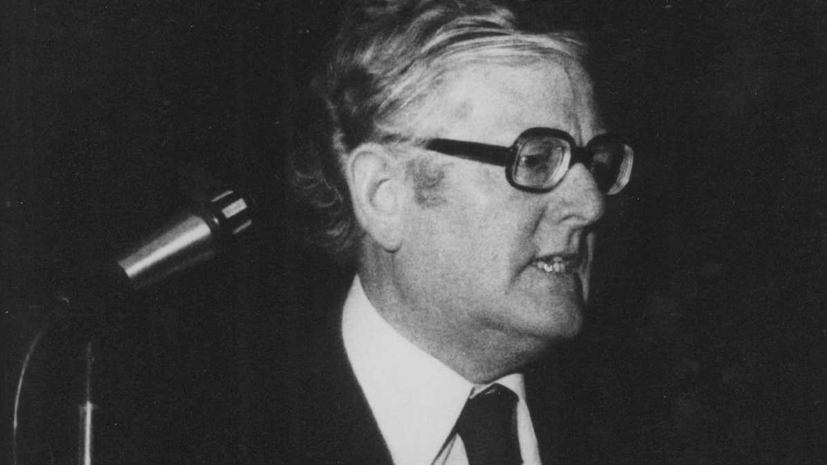 Old black and white, slightly grainy photo of man in suit and tie giving a speech