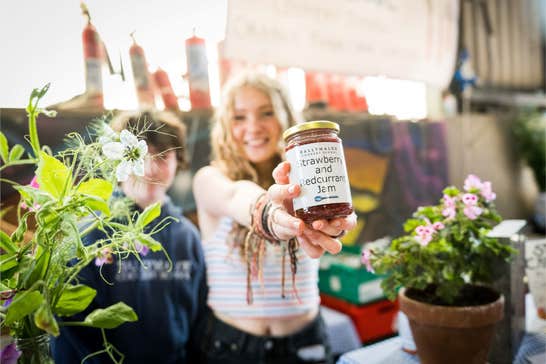 A huge selection of craft food makers, a woman is holding up a jar of jam towards the camera, smiling.