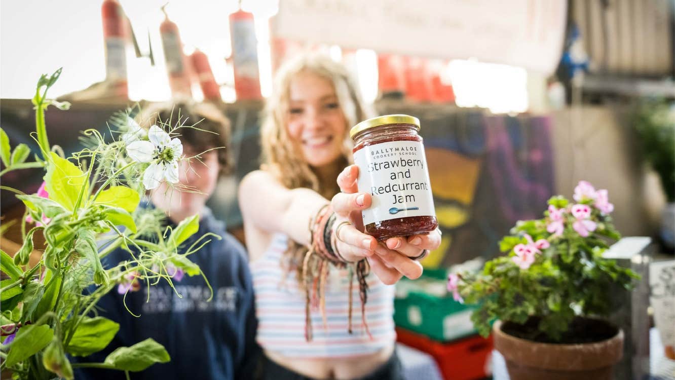 A huge selection of craft food makers, a woman is holding up a jar of jam towards the camera, smiling.