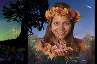 A smiling woman with headband made of yellow and orange flowers with palms outstretched holding a small, cartoon image of a donkey wearing red clothes, bordered with flowers and black silhouette of tree with small green fairy underneath.
