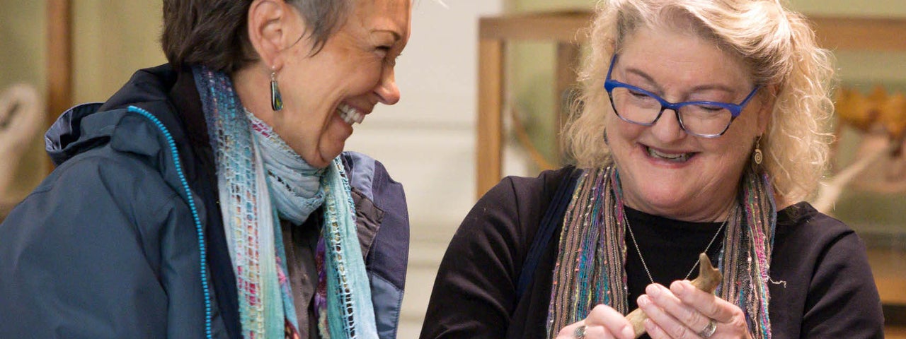 2 women laughing, both holding small objects, display cabinets in the background.