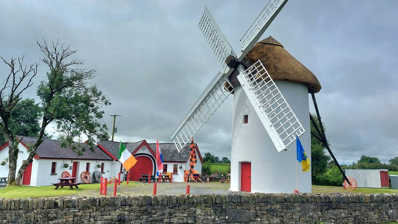 The exterior of the restored Elphin Windmill with a thatched roof and red door and the agricultural museum in the background