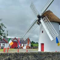 The exterior of the restored Elphin Windmill with a thatched roof and red door and the agricultural museum in the background