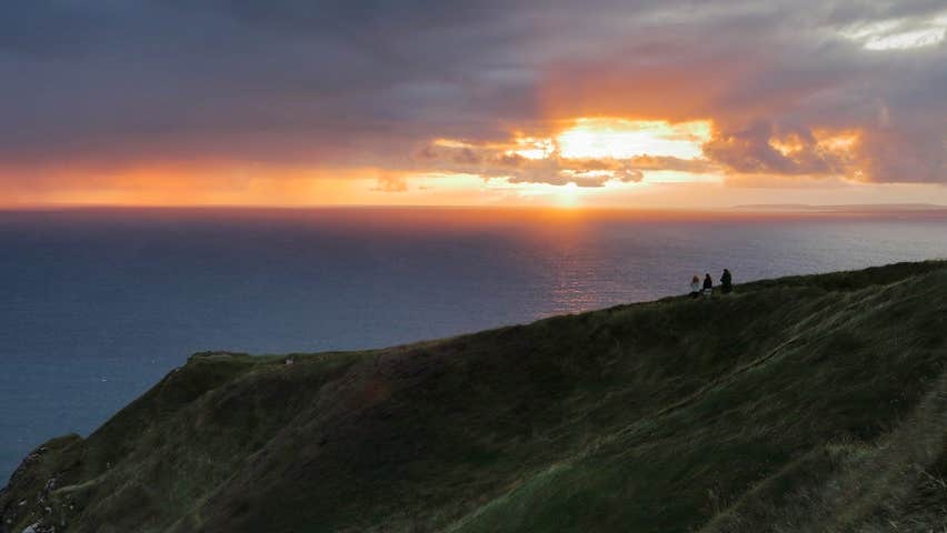 People stand on a cliff area looking out at a sunset