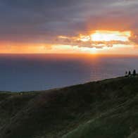People stand on a cliff area looking out at a sunset