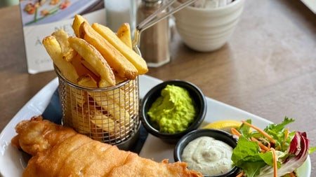 Basket of chips with a piece of battered fish and sides on a while rectangular plate