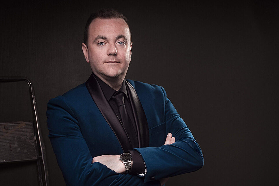 Country and Gospel singer, Patrick Feeney pictured in dark colours with arms crossed with black background