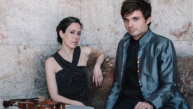 A woman in a black, sleeveless dress is seated beside a man in a dark grey shiny jacket with a violin lying on a small table in front of them against a tall grey stone background.